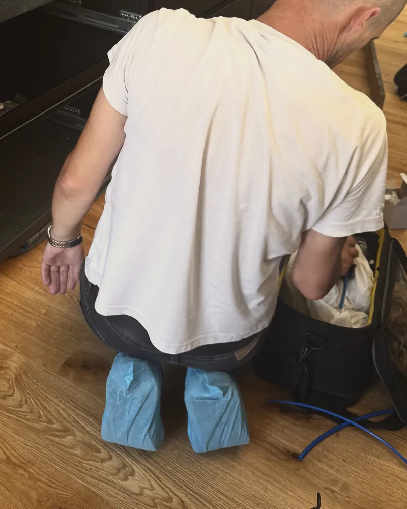 Un installatore di spalle, accovacciato con i copriscarpe azzurri, accanto alla borsa degli attrezzi