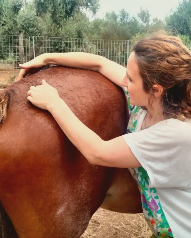 Una donna con i capelli ricci appoggia le mani e l’avambraccio sul dorso di un cavallo baio, all’aperto